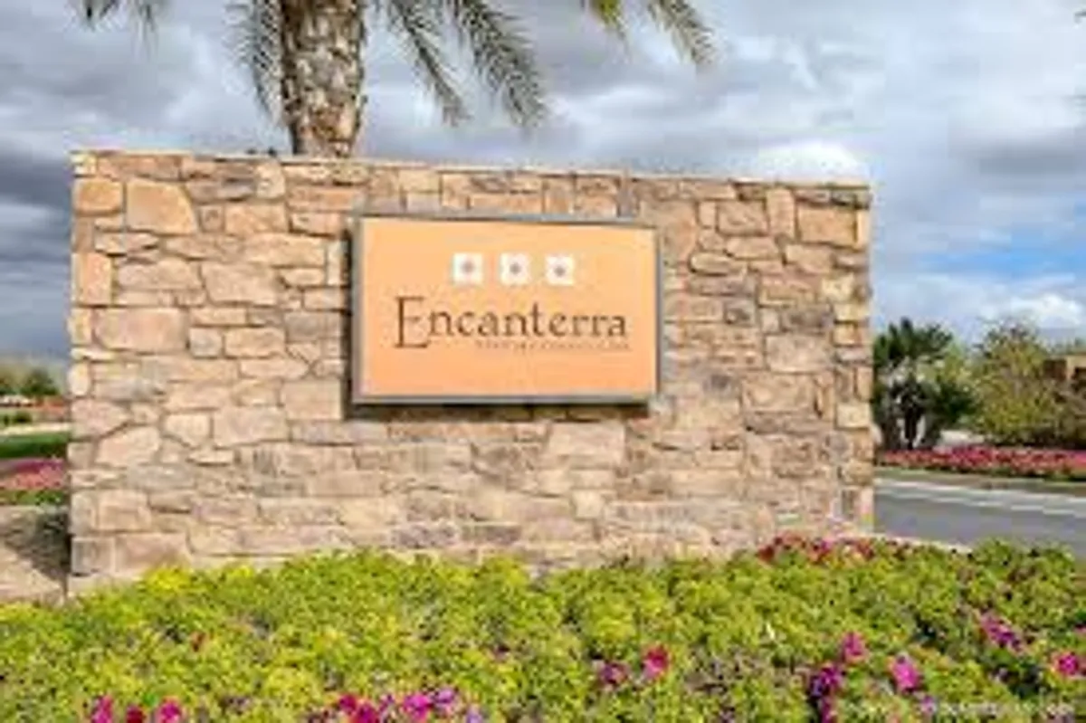 A beautifully landscaped entrance sign framed by vibrant flower beds and tall palm trees, signaling a welcoming arrival into the community. The stone sign suggests a sense of place and community pride, while the lush planting and clear skies evoke the inviting outdoors.