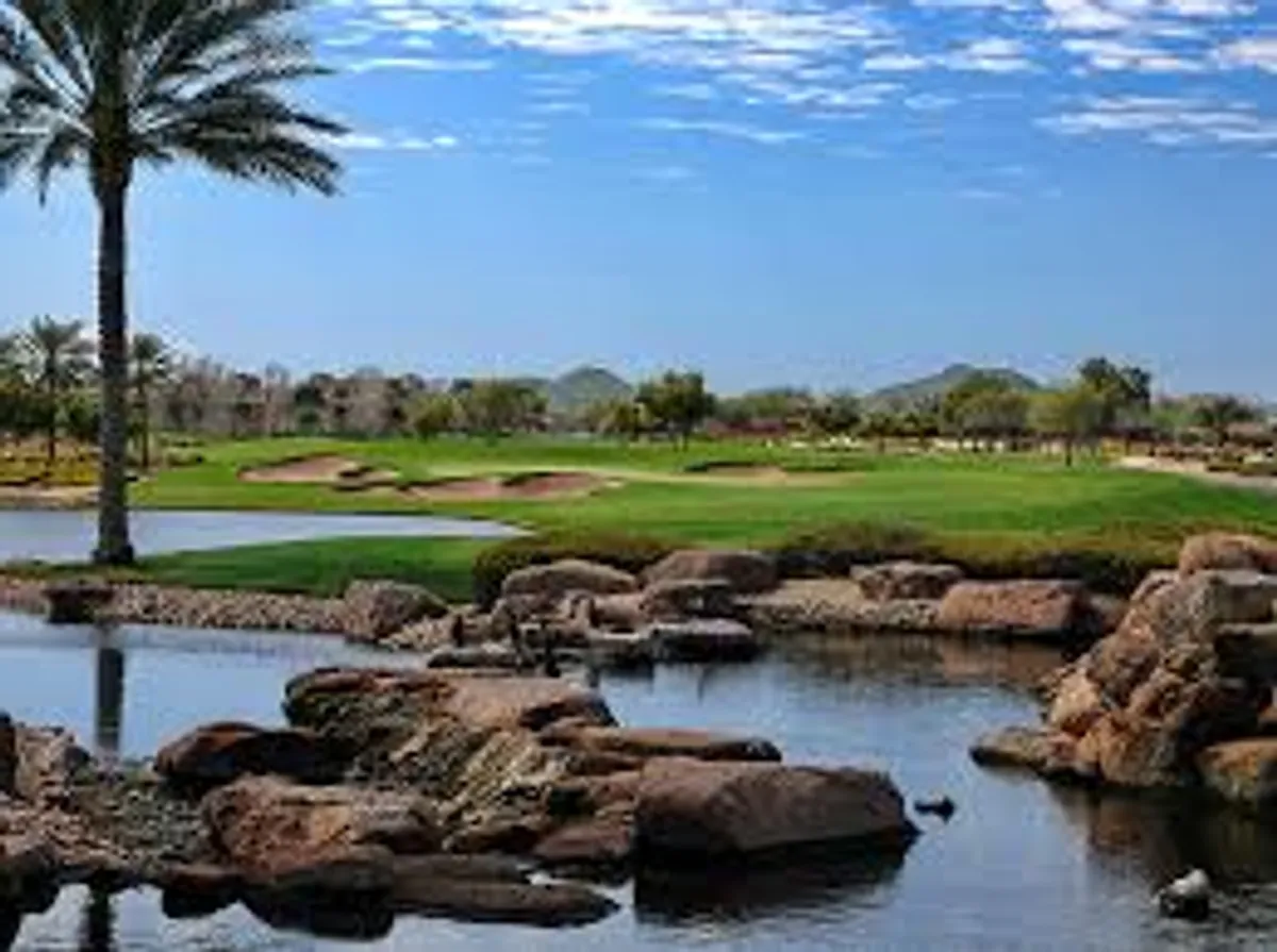 This image presents a sweeping view of the golf course at Encanterra featuring lush green fairways, a tranquil water feature, palm trees, and striking rock formations, all under an expansive sky. The scene captures the serenity and challenge of championship-level golf while also hinting at the broader natural beauty residents can enjoy.