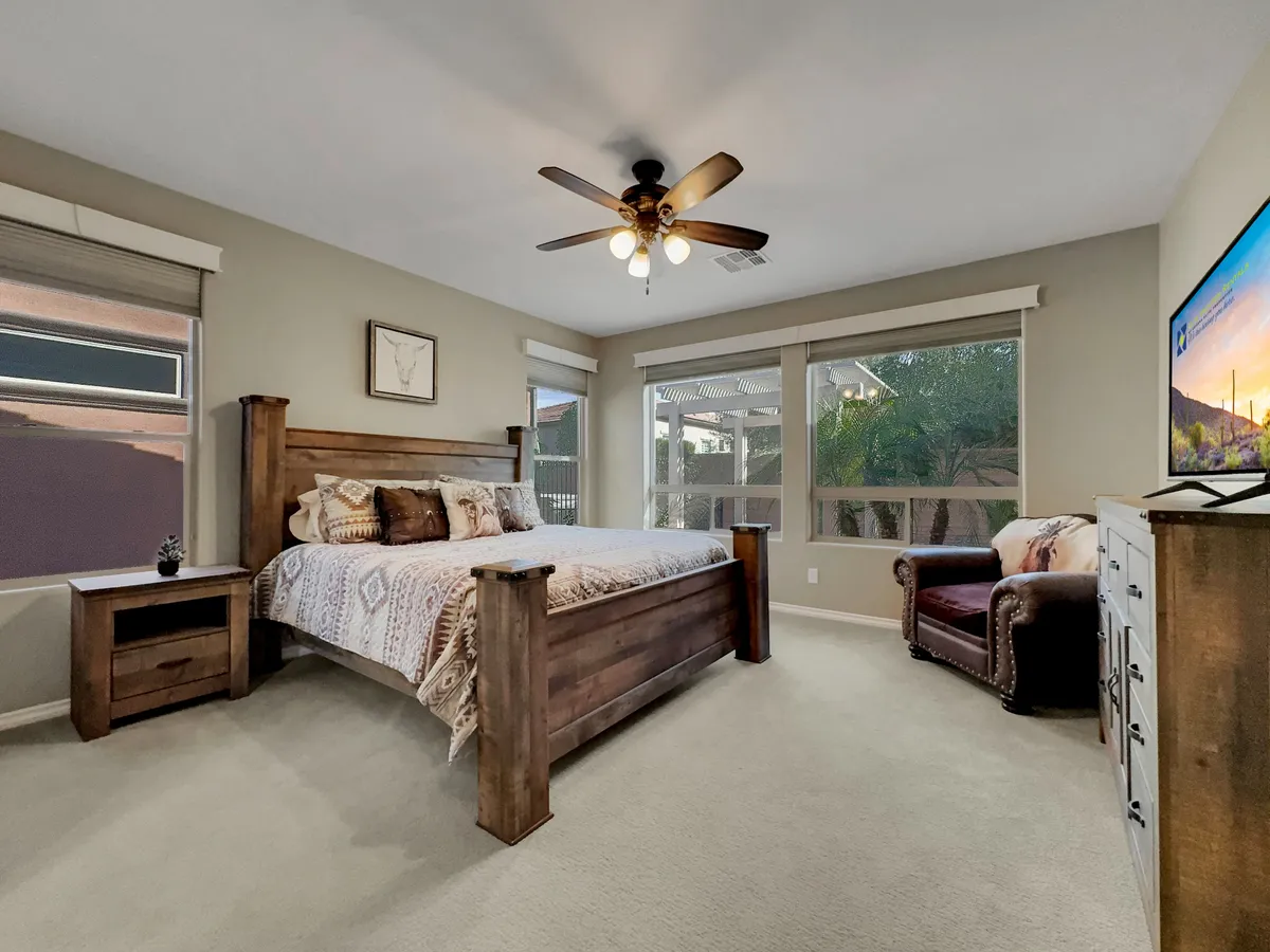 A tranquil primary bedroom featuring a wooden bed frame and a comfortable armchair set beside large windows. Ambient natural light streams in, highlighting the decorative wall art and creating a restful atmosphere perfect for relaxing, reading, or enjoying a morning coffee. The space feels both spacious and private, with amenities such as a TV visible for added comfort.