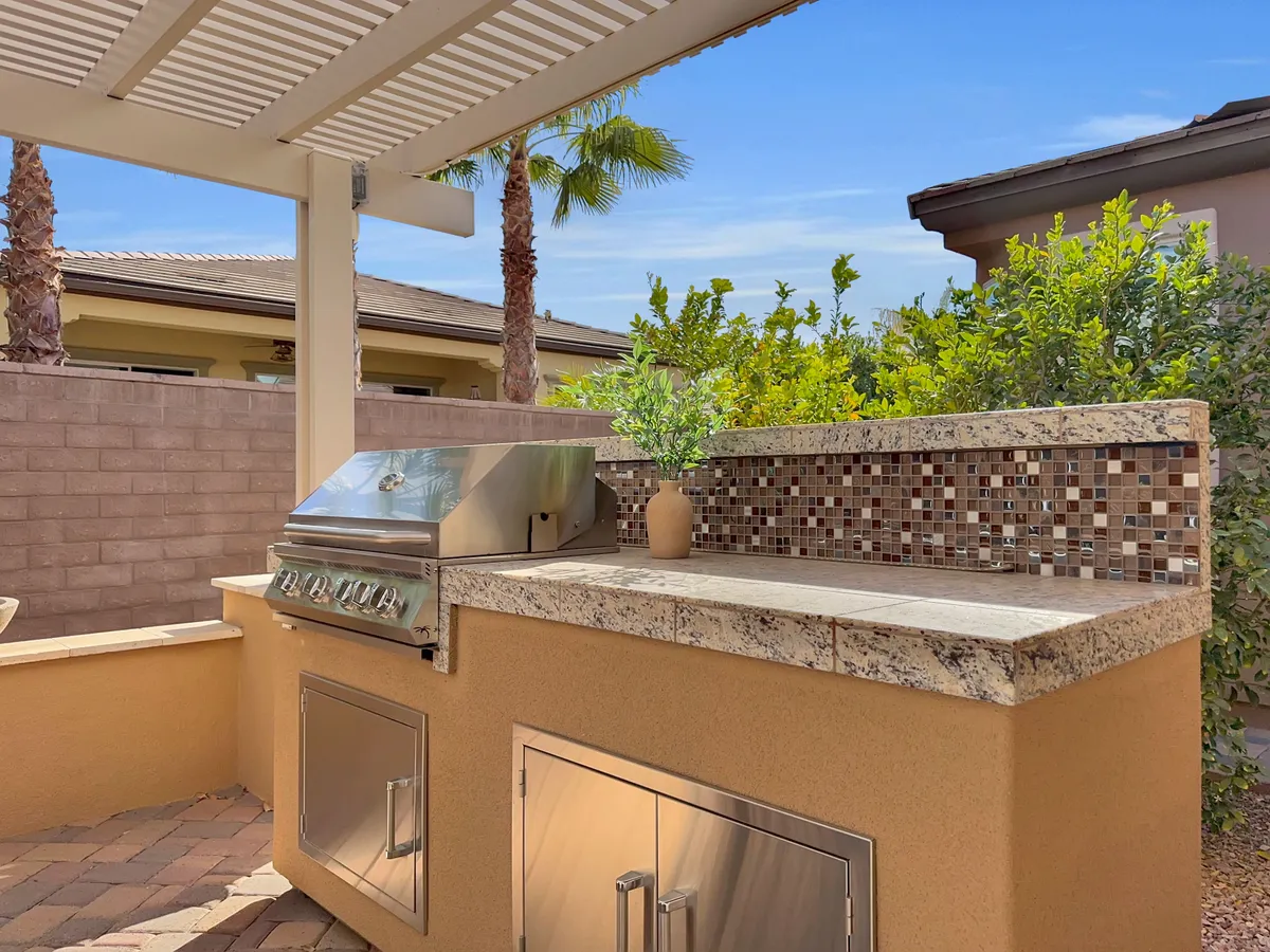 An exterior view of a well-appointed outdoor grilling area with granite countertops, a mosaic backsplash, and potted plants. The scene is set up for casual outdoor entertaining, suggesting vibrant social gatherings and community barbecues.