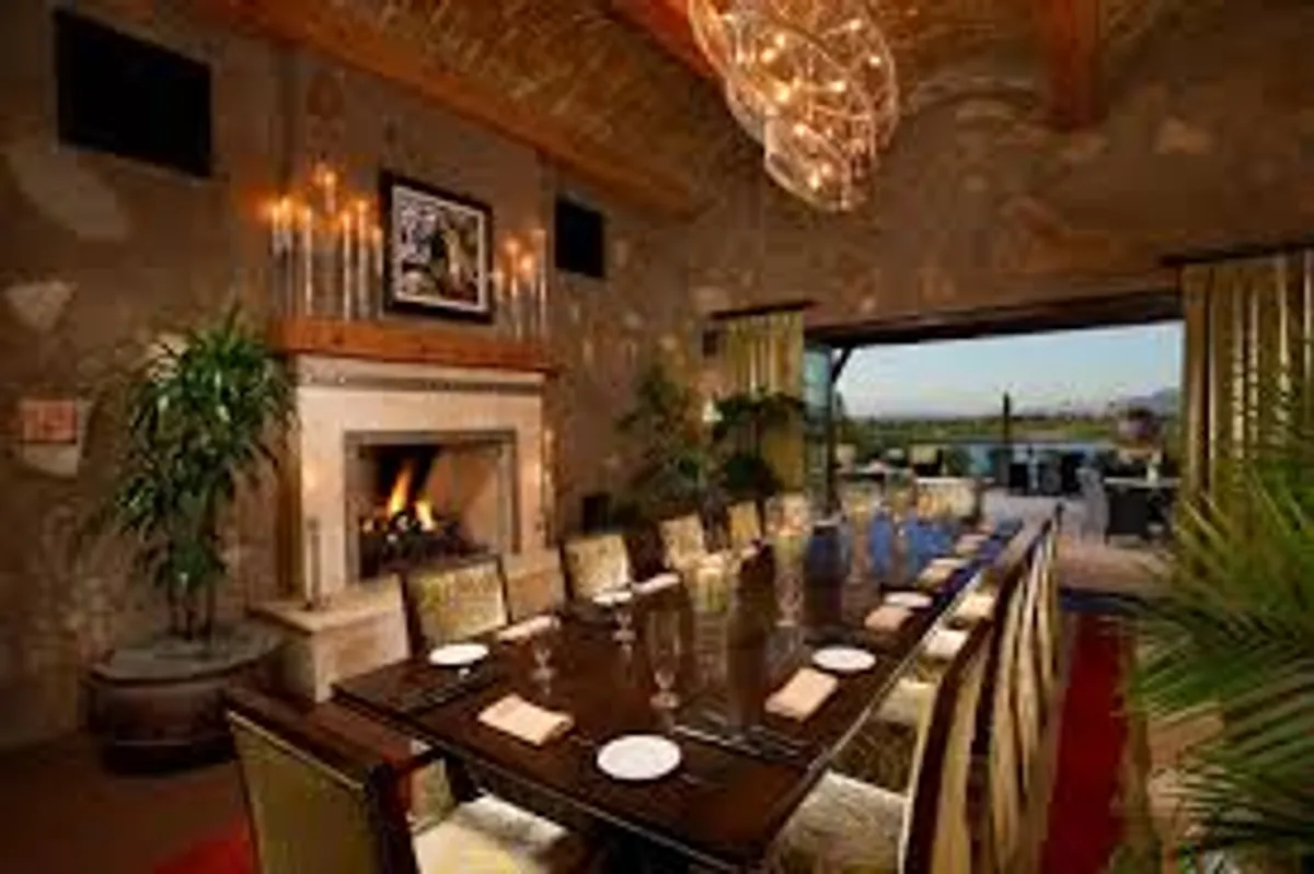 A spacious dining area opens to the outdoors via large open doors, with a chandelier above a large table and a cozy fireplace nearby. The view blends the comfort of the interior with seamless access to a scenic outdoor patio, perfect for social gatherings and relaxation.