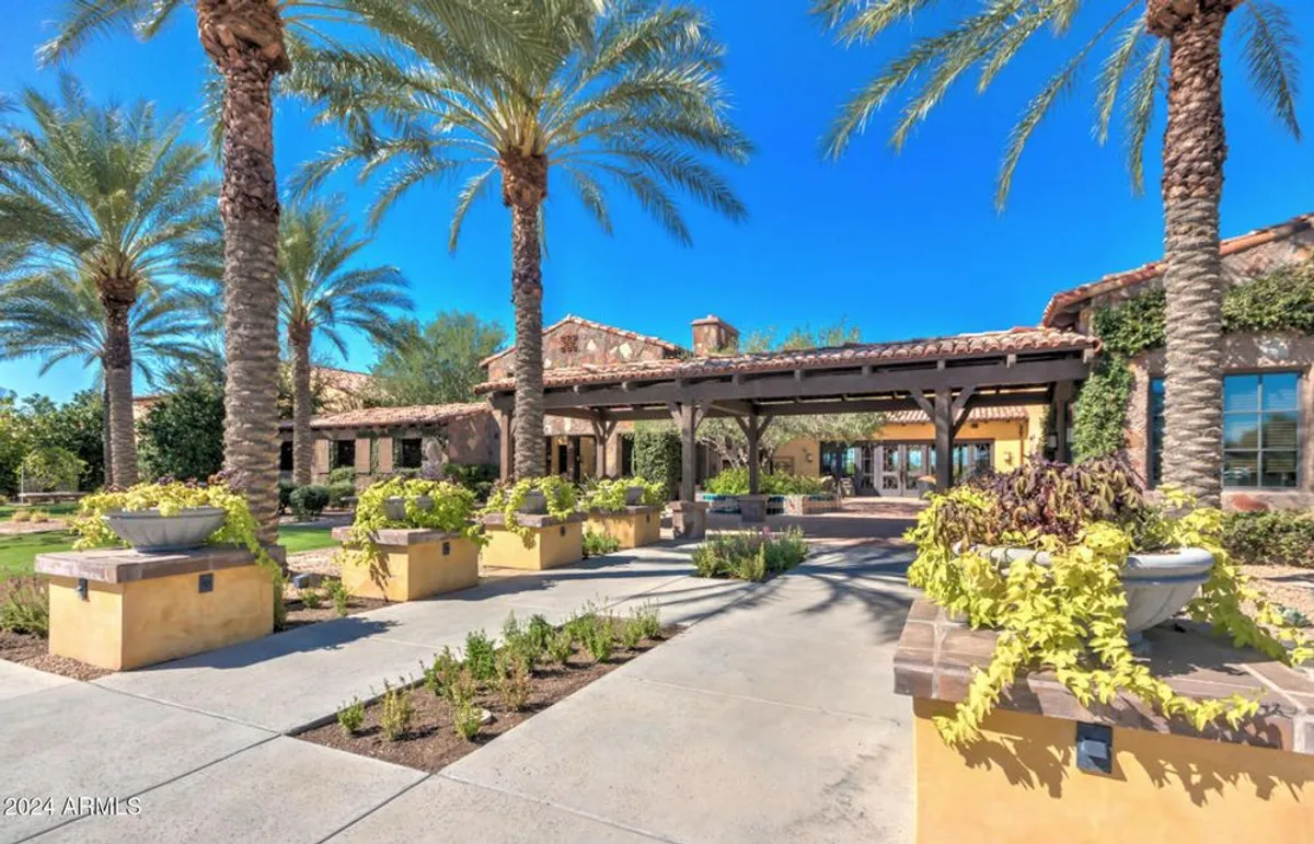 This exterior image showcases a lush garden courtyard framed by palm trees, a welcoming entrance archway, and vibrant flower pots. The warm glow of sunset filters through, highlighting the serene outdoor ambiance and the sense of arrival.