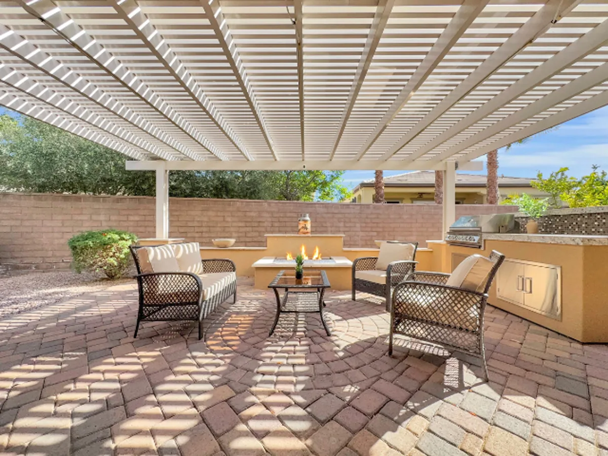 This image showcases a stylish outdoor seating area under a pergola, centered around a modern fire pit and adjacent to a barbecue grill. The surrounding lush greenery and soft lighting create a cozy, inviting setting perfect for relaxing or entertaining guests.